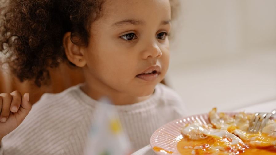 Charming young girl with curly hair enjoying a meal inside at a casual setting.