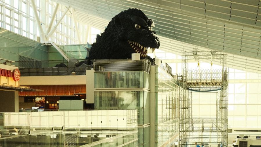 Iconic Godzilla statue inside a bustling, modern airport terminal with a high ceiling.