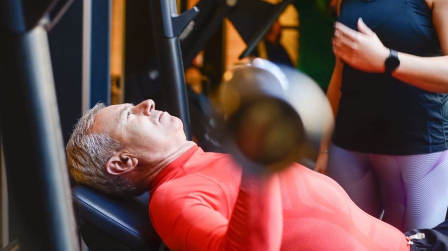 Elderly man exercising with a barbell at the gym under supervision.