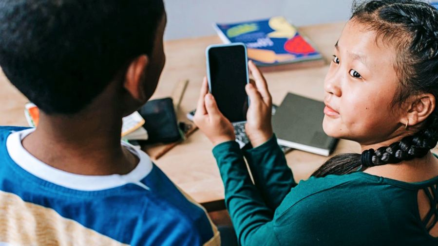Two children using a smartphone at a classroom desk, engaging in study.