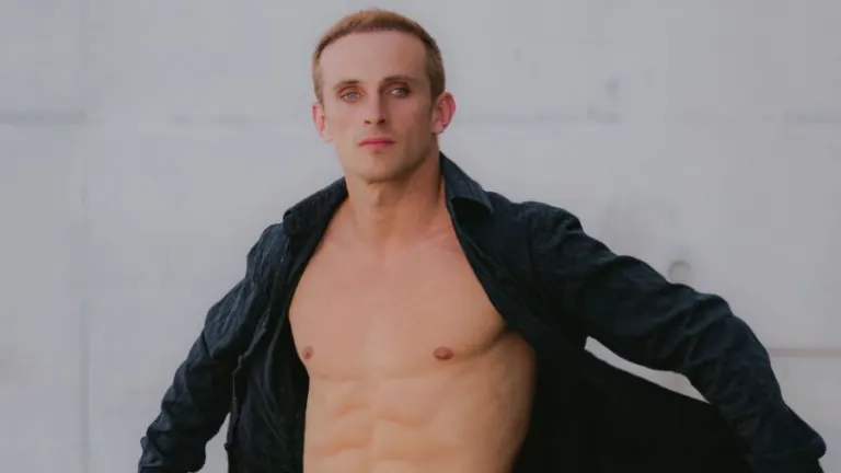 Athletic shirtless man in black jacket standing confidently against urban wall.