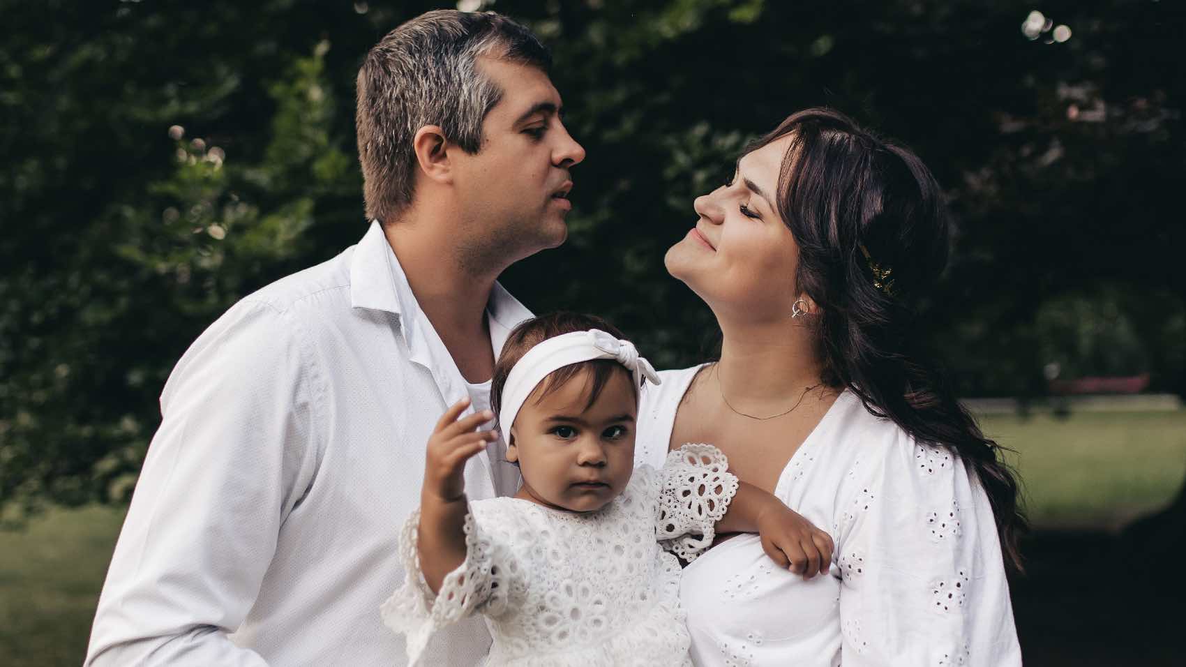 A happy family with parents holding their baby daughter in a sunny park setting.