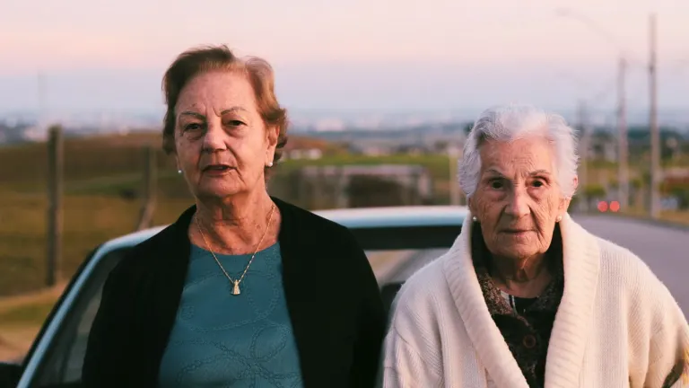 Two senior women standing together outdoors by a car during sunset.