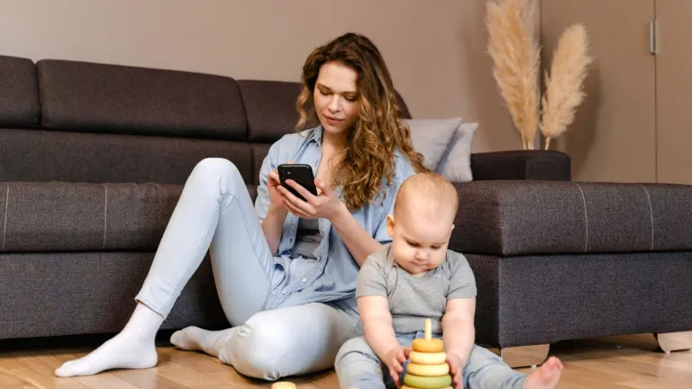 A young mother sits with her toddler playing on the floor, focusing on her phone.