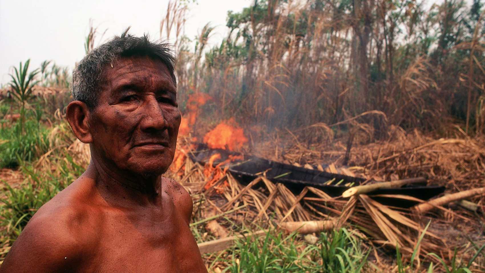 Shirtless Indigenous man standing by a small fire in a dry, grassy wilderness setting.