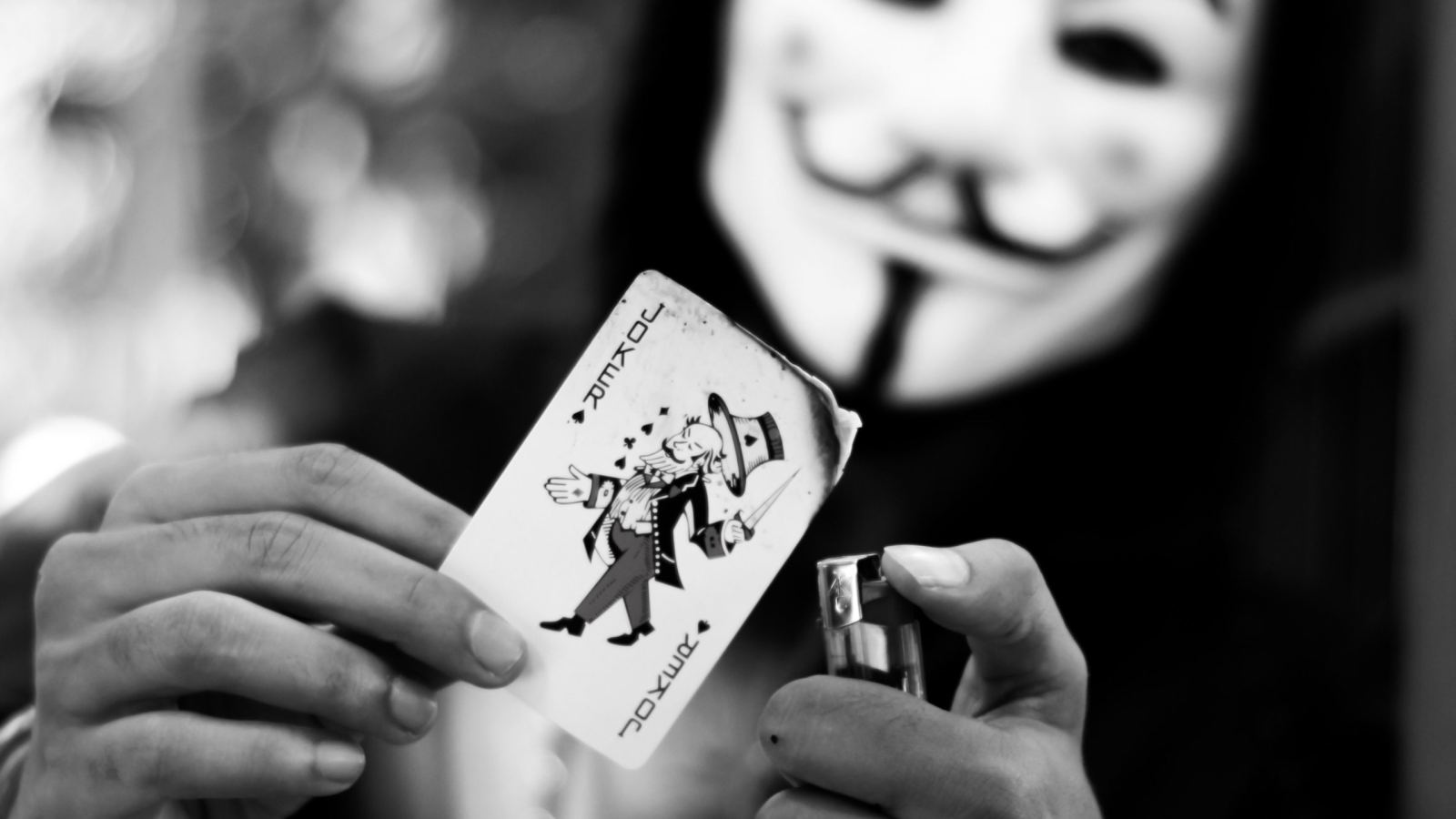 Black and white image of a masked figure holding a Joker card near a lighter flame.