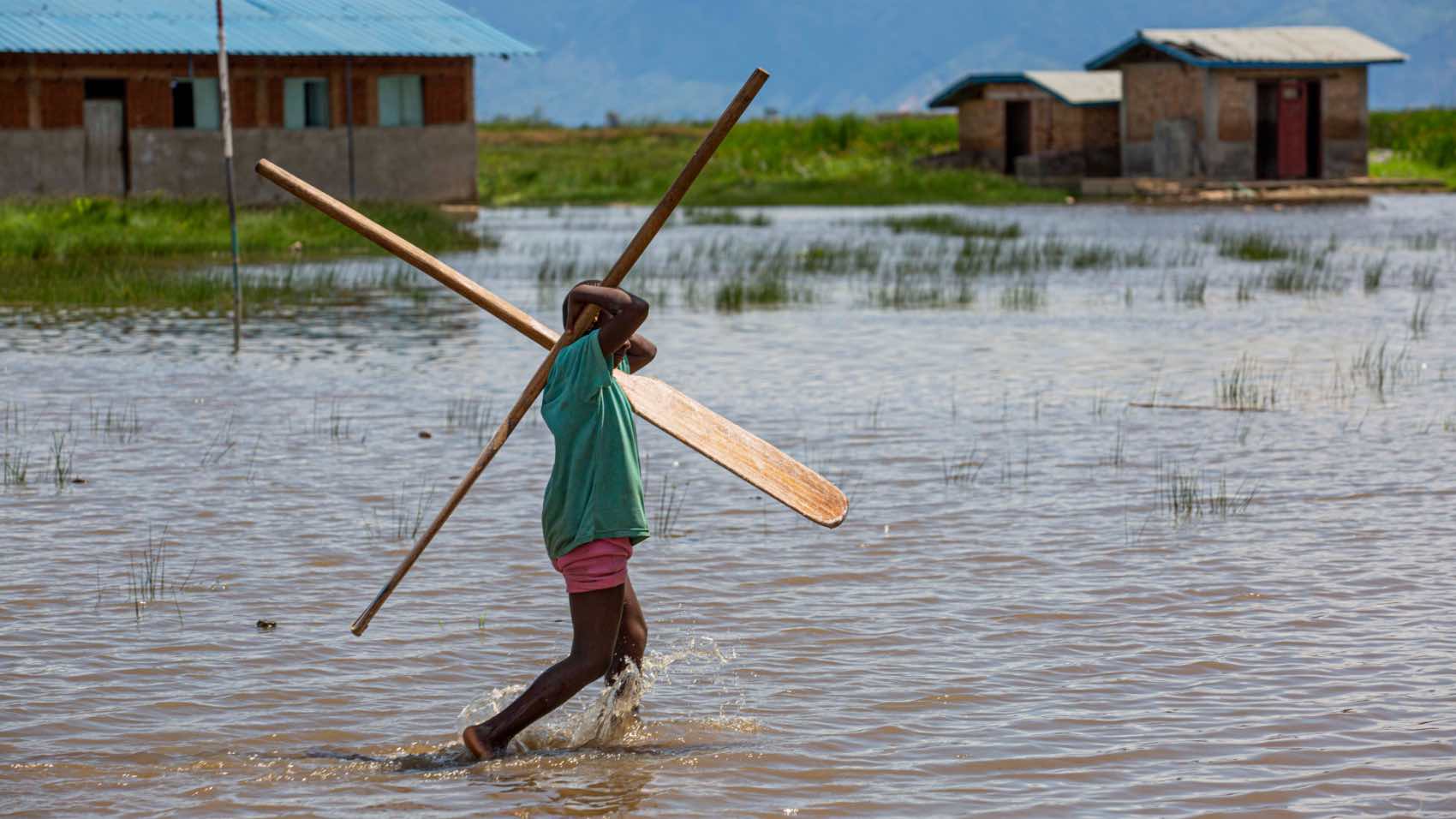 A boy walks through water carrying canoe paddles near rustic houses on a flooded plain.