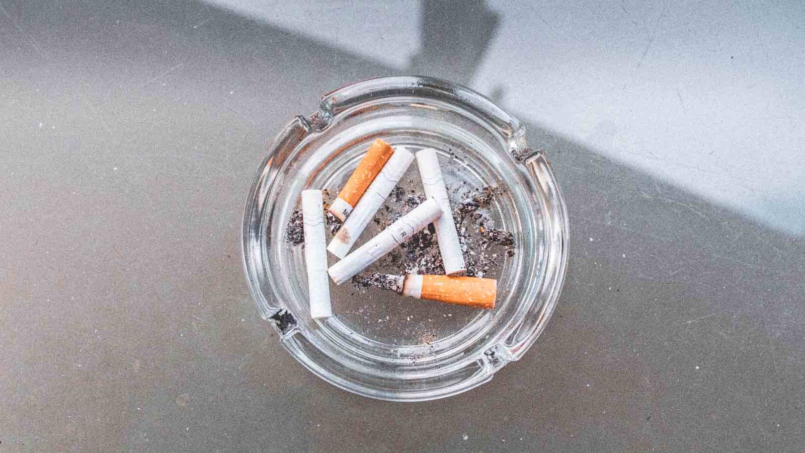 Glass ashtray with used cigarettes on a glass table in an outdoor setting.
