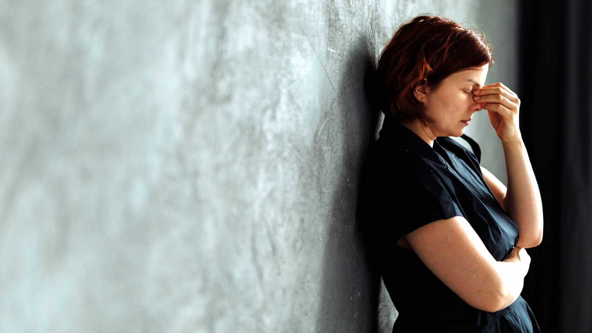 A woman in stress with eyes closed, leaning against a concrete wall indoors.