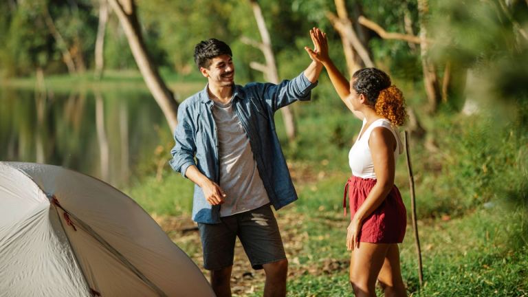 A couple enjoying a summer camping trip by a lake, sharing a high-five next to a tent.