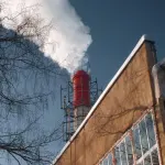 A factory chimney emits white smoke, casting a shadow on a clear day beside a bare winter tree.