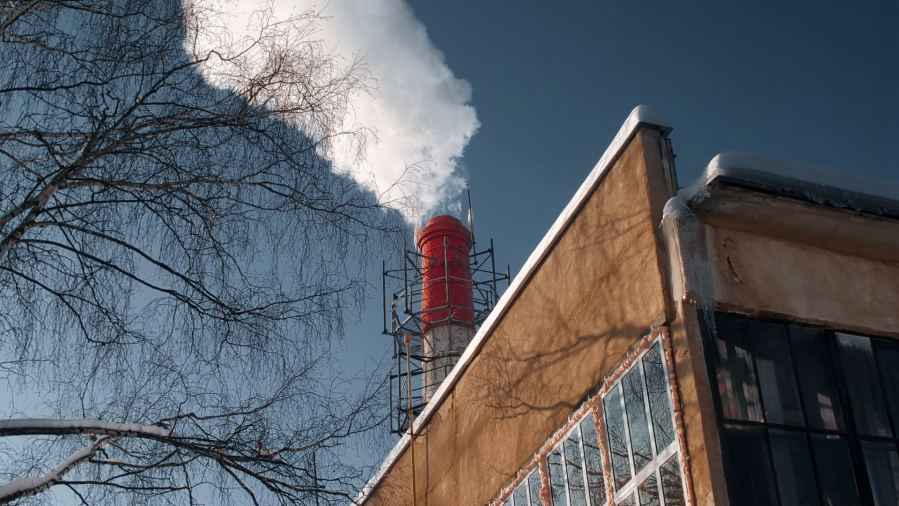 A factory chimney emits white smoke, casting a shadow on a clear day beside a bare winter tree.