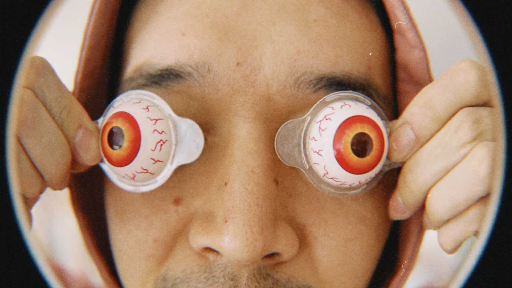 Creative portrait of a man in a hoodie holding toy eyeballs over his eyes, creating a whimsical effect.