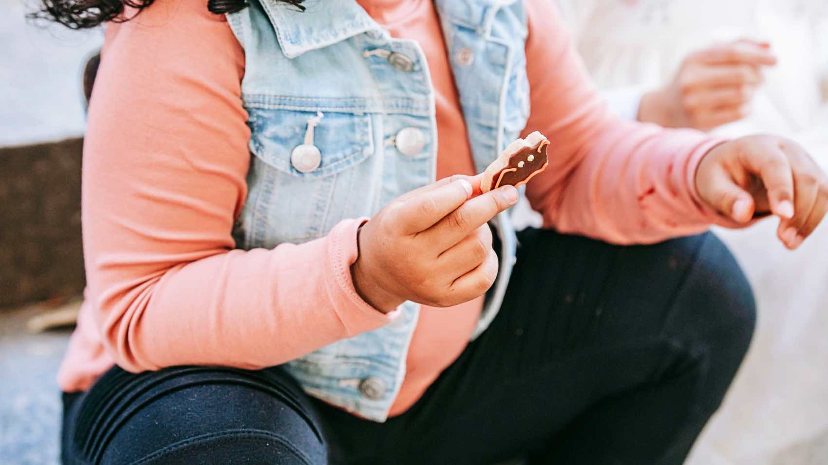 Close-up of a young child holding a Halloween-themed gingerbread cookie while sitting outdoors in autumn attire.
