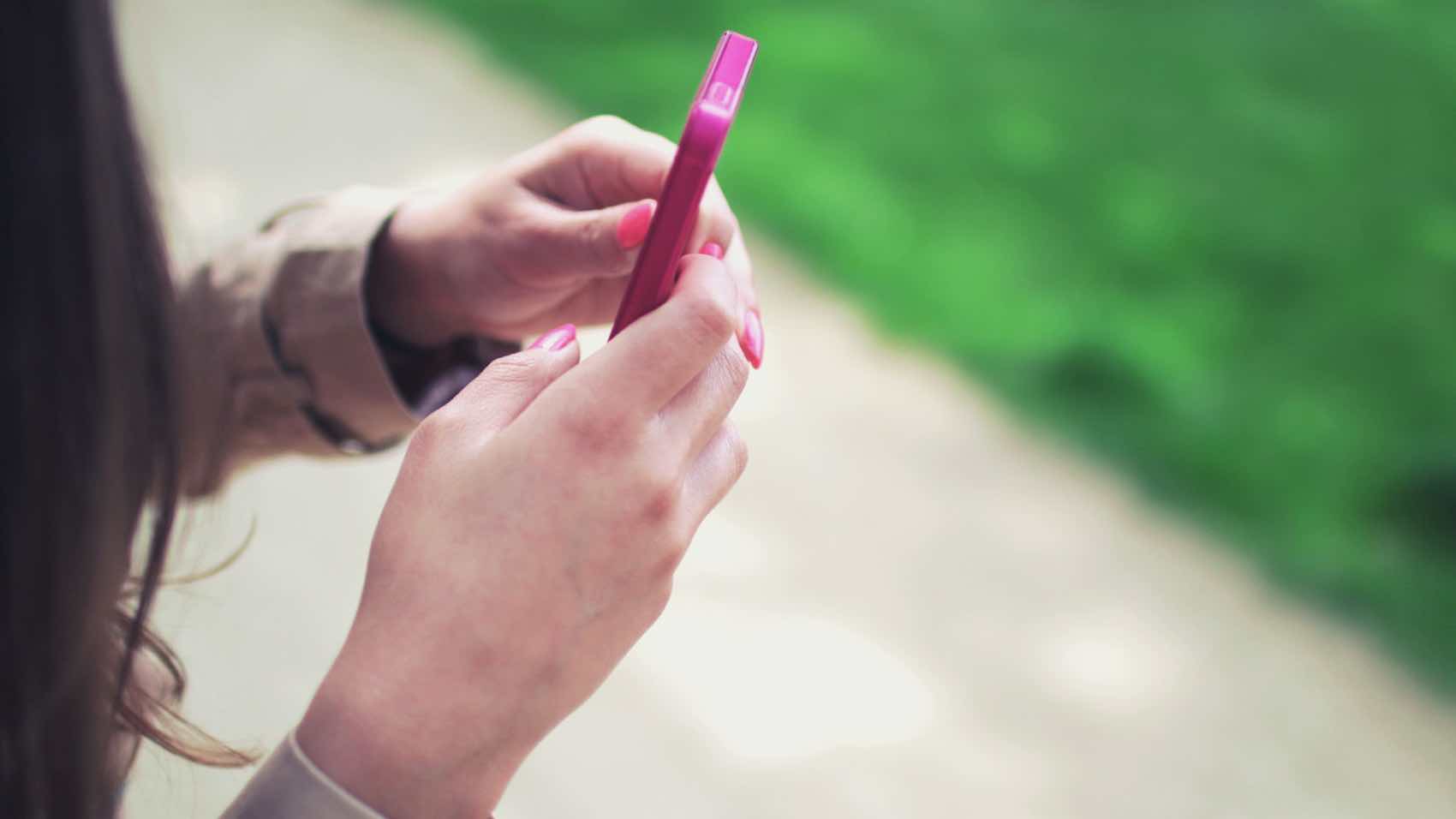 Woman holding a pink smartphone with both hands in a casual outdoor setting.