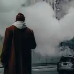 A man in a brown coat stands in a smoky city street surrounded by tall buildings.
