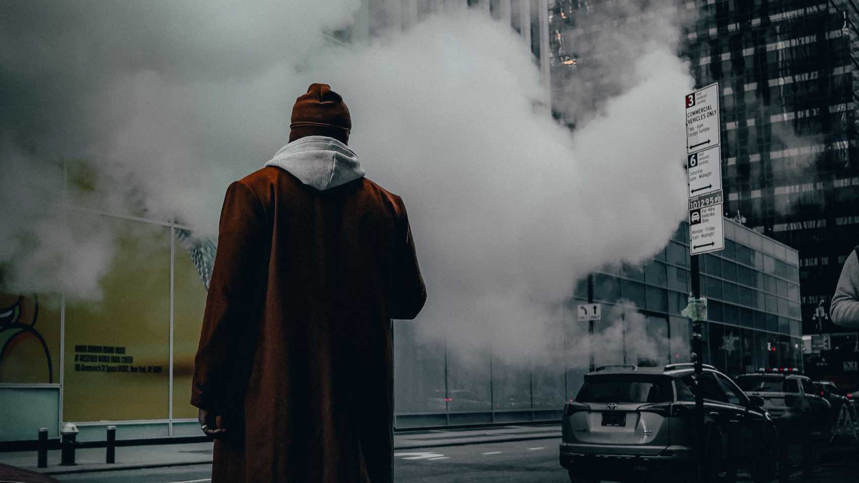 A man in a brown coat stands in a smoky city street surrounded by tall buildings.