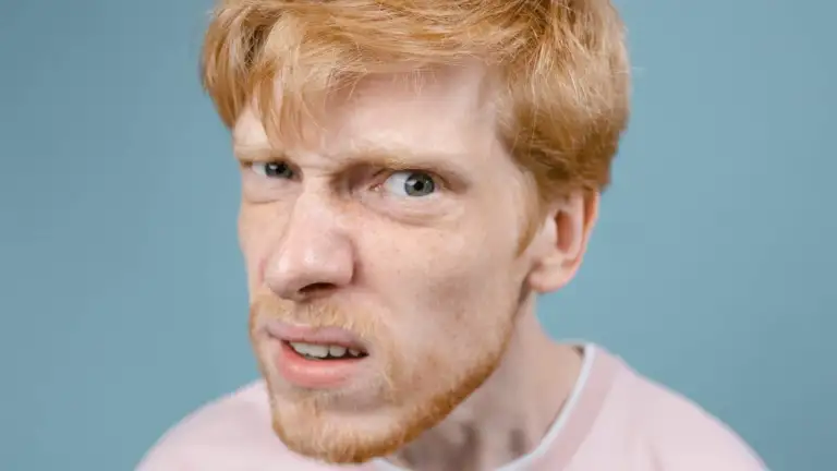 Redhead man with a comical expression wearing a pink shirt against a blue background.