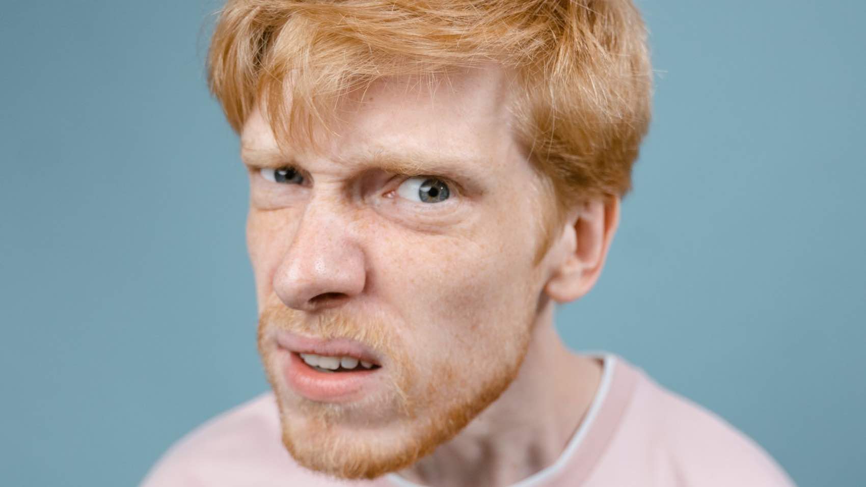 Redhead man with a comical expression wearing a pink shirt against a blue background.