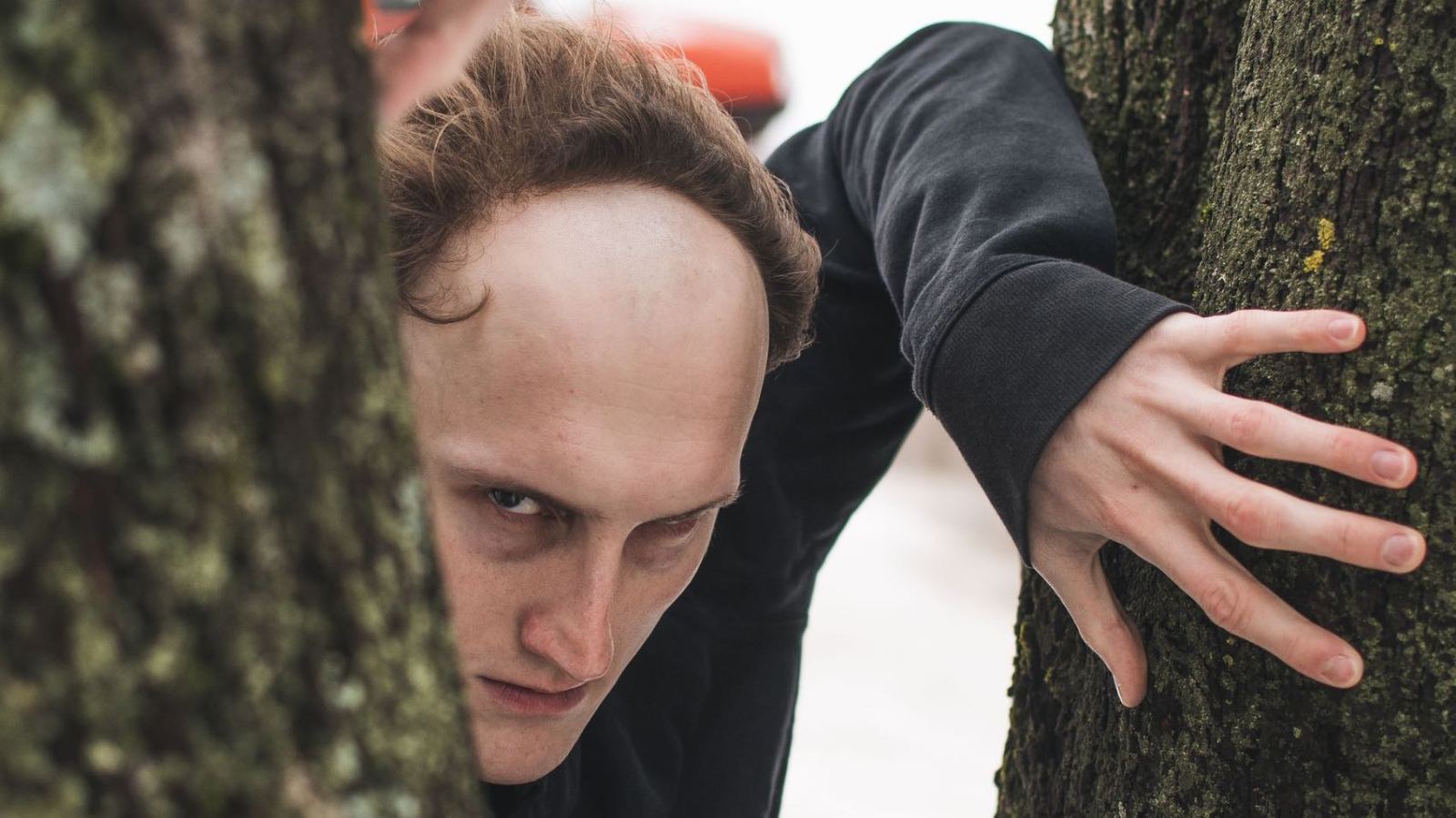 Eerie portrait of a man in a black jacket peering through tree trunks.