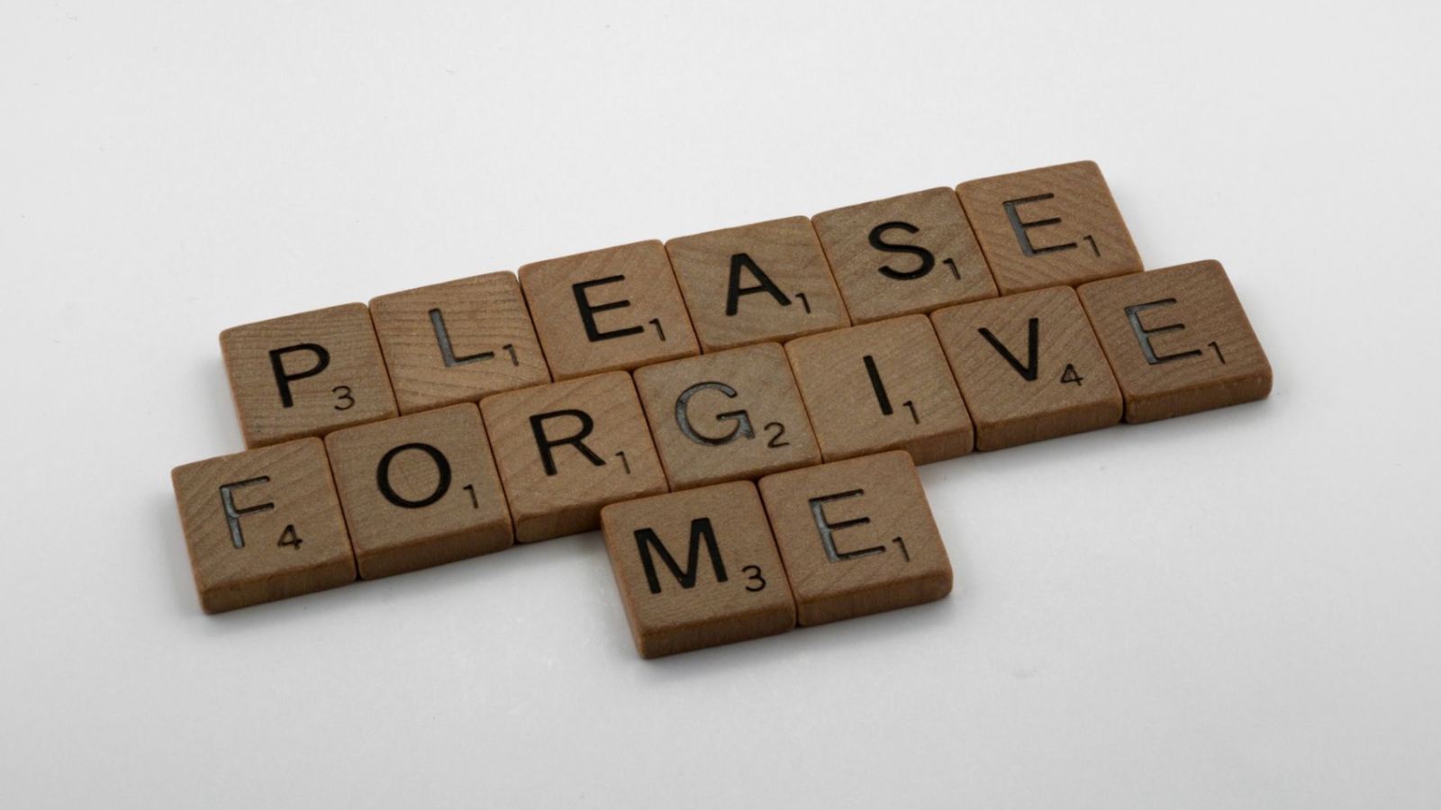 Wooden Scrabble tiles spelling 'Please Forgive Me' on a white surface, emphasizing forgiveness.