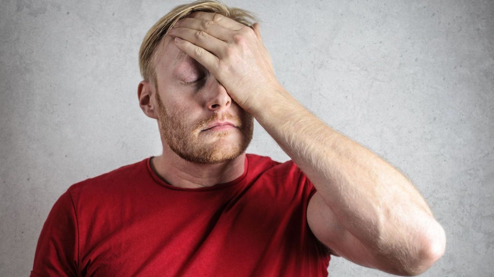 A frustrated man in a red shirt holds his head in stress against a neutral background.