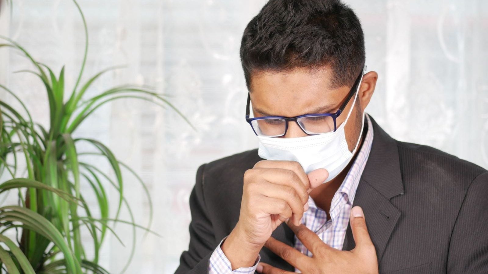 Asian man coughing in a black suit with eyeglasses and face mask indoors.